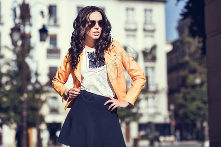 带太阳镜的轻黑发女人女孩,时尚模特,穿着橙色现代夹克蓝色裙子,站城市背景图片