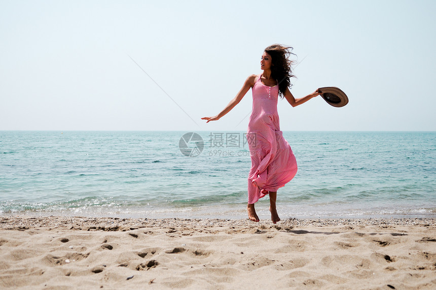 位穿着粉红色长裙的美女热带海滩上跳跃的肖像图片