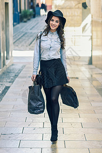 美丽的黑发轻女子穿着短裙牛仔衬衫走街上,带着购物袋图片