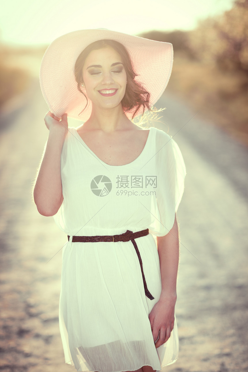 春天的条乡村道路上闭着眼睛的轻女人的肖像穿着白色连衣裙,棕色腰带粉红色太阳帽的女孩背光摄影与太阳女人的后图片