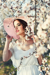 黑发花的春天花田里轻女人的肖像杏仁花开花了穿着白色连衣裙粉红色太阳帽的女孩背景
