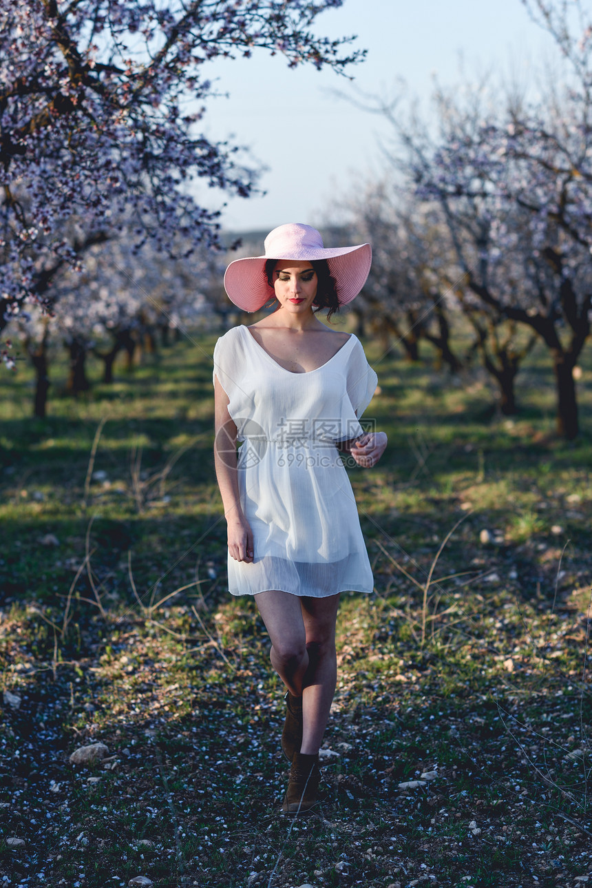春天花田里轻女人的肖像杏仁花开花了穿着白色连衣裙粉红色太阳帽的女孩图片