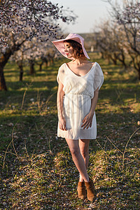 黑发花的春天花田里轻女人的肖像杏仁花开花了穿着白色连衣裙粉红色太阳帽的女孩背景