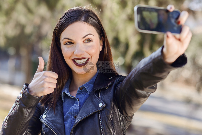 公园里个漂亮的轻女人用智能手机自拍的肖像图片