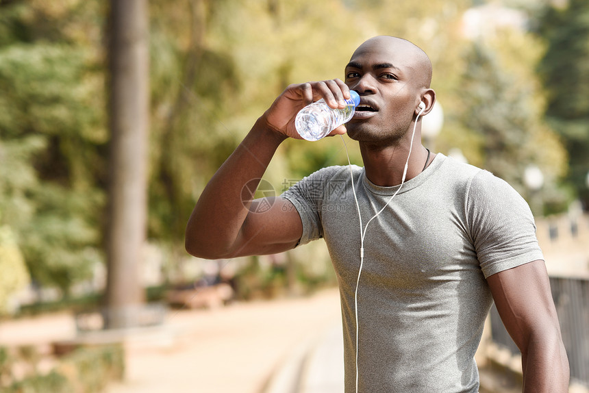 轻的黑人城市背景下跑步前喝水轻的男用赤裸的躯干锻炼,用耳机听音乐图片