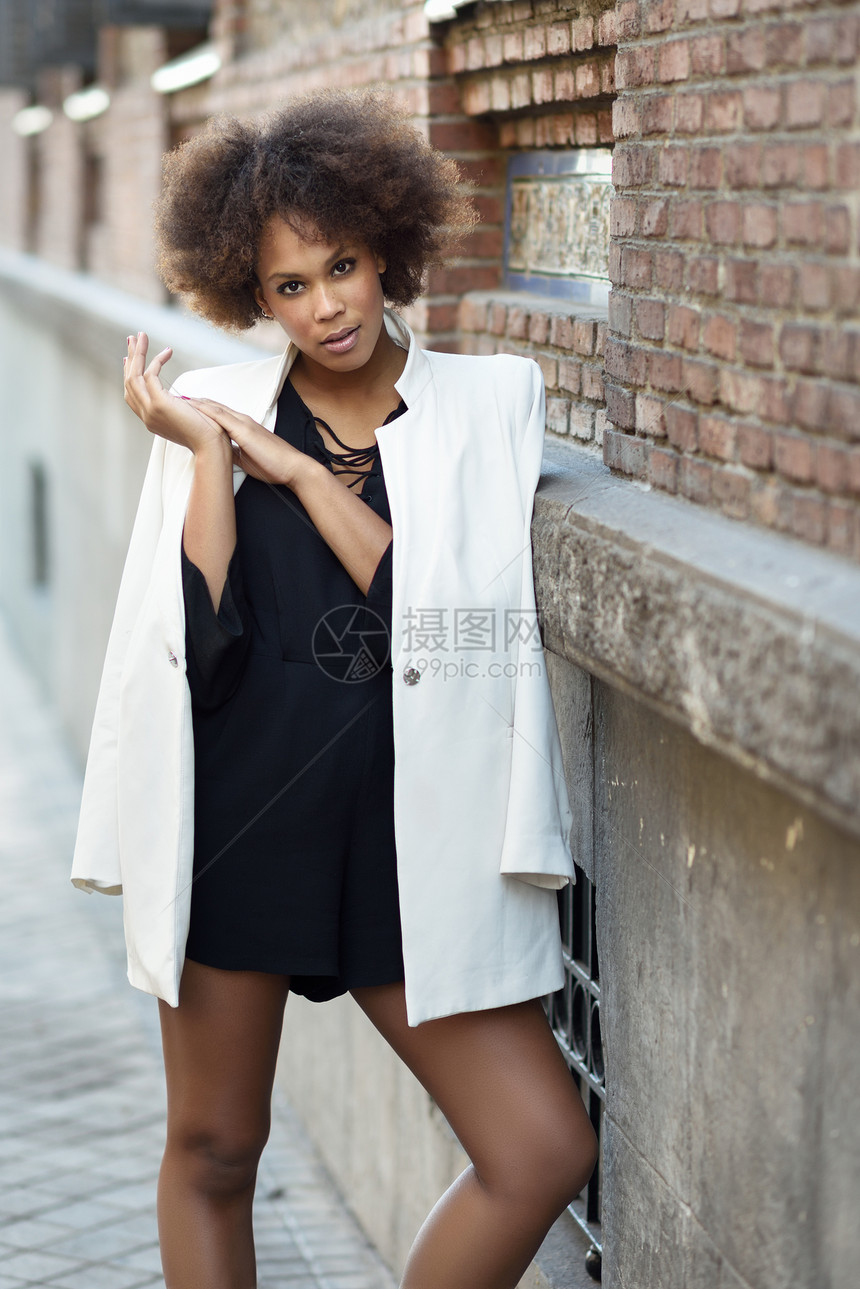 轻的黑人女,留着AFRO发型,站城市背景中穿着白色夹克黑色连衣裙的混合女孩砖墙附近摆姿势图片