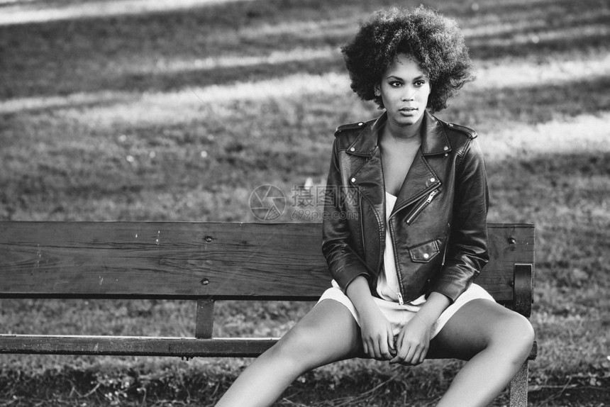 轻的黑人女,留着AFRO发型,坐城市公园的长凳上混合女人穿红色皮夹克白色连衣裙与城市背景图片