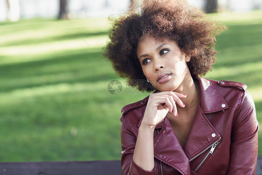 轻的黑人女,留着AFRO发型,坐城市公园的长凳上混合女人穿红色皮夹克白色连衣裙与城市背景图片