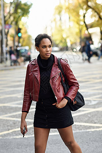 轻的黑人女,留着AFRO发型,站城市街道上混合女人穿着红色皮夹克,黑色毛衣城市背景的裙子女携带袋智能手机背景图片