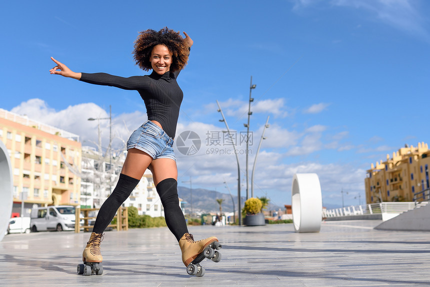 轻合身的黑人妇女溜冰鞋骑户外的城市街道上张开双臂阳光明媚的日子里,微笑的女孩留着AFRO的发型图片