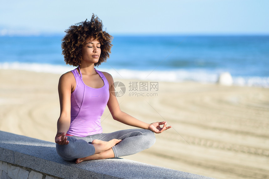 黑色女人,阿弗罗发型,海滩上瑜伽,闭着眼睛轻的女穿着运动服,以莲花的姿态,背景分散图片