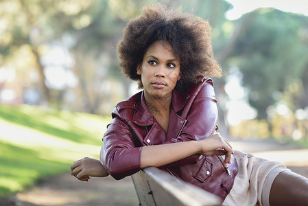 轻的黑人女,留着AFRO发型,坐城市公园的长凳上混合女人穿红色皮夹克白色连衣裙与城市背景背景图片