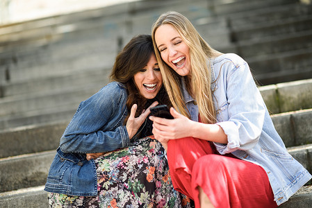 两个女孩户外看着智能手机上的些趣的视频,坐城市的台阶上女朋友女孩笑图片