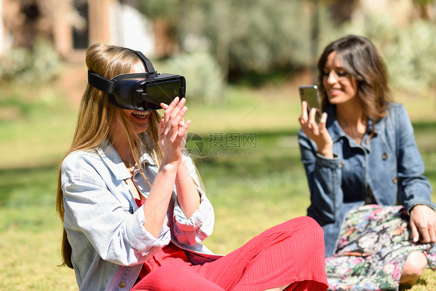 女人看着VR眼镜,双手户外手势,城市公园,她的女朋友用智能手机拍了段视频图片