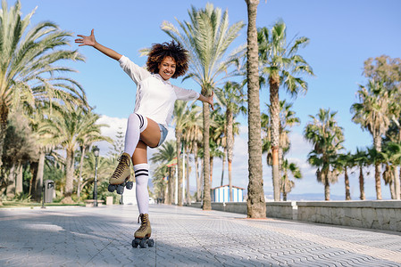 留音机微笑的黑人妇女溜冰鞋骑户外海滩长廊与棕榈树阳光明媚的日子里,微笑的女孩留着AFRO的发型背景
