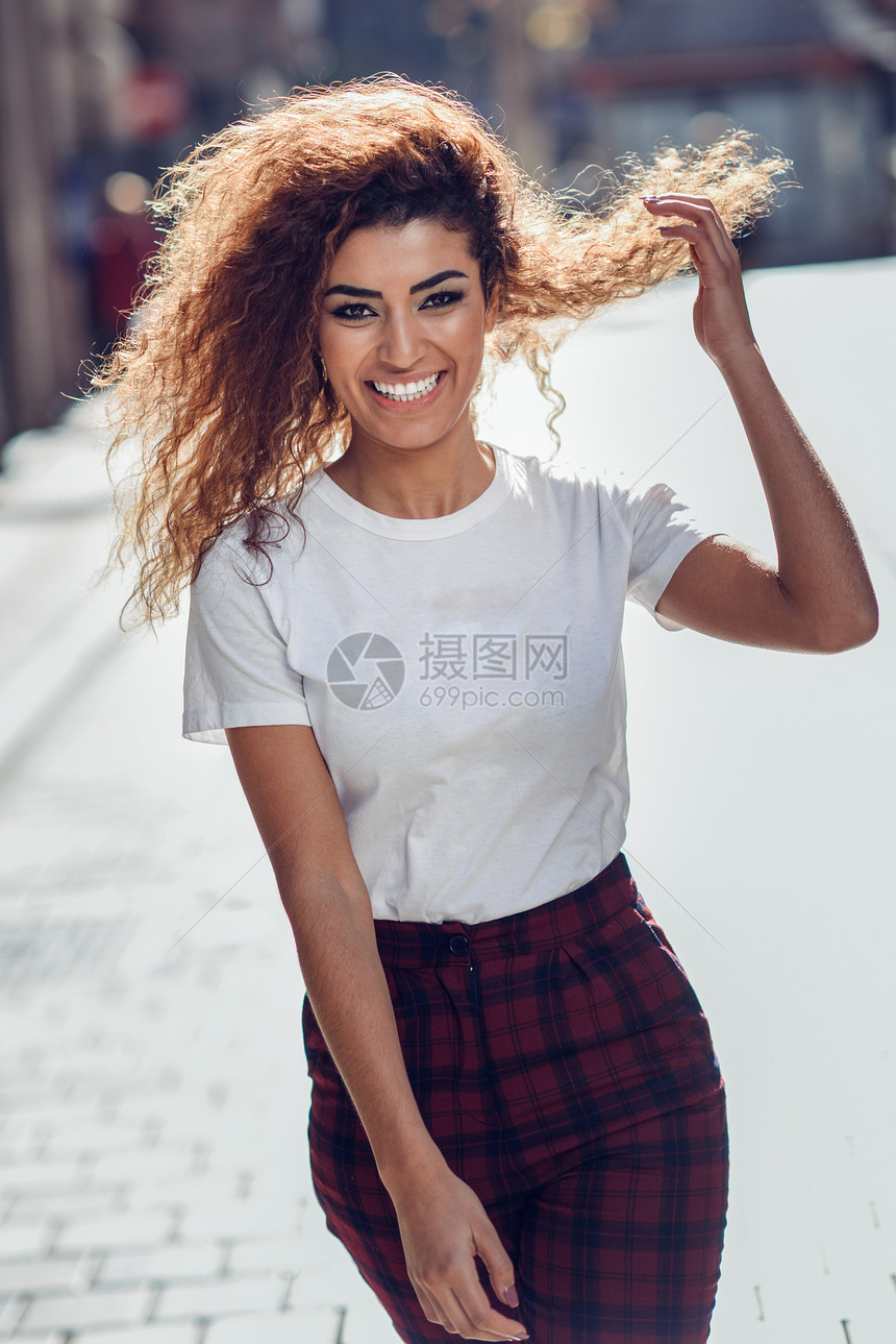 微笑的轻阿拉伯女人,黑色卷发街上穿便装的阿拉伯女孩快乐的女穿着白色T恤检查裤子图片