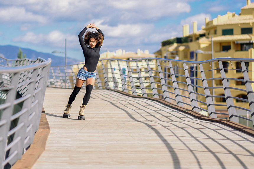 阿夫罗式发型女人溜冰鞋骑户外的城市B轻的适合黑人妇女溜冰鞋骑户外的城市桥上张开双臂阳光明媚的日子里,微笑的女孩留图片