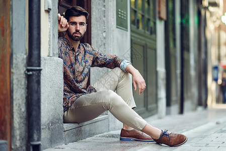 轻的留着胡子的男人,时尚的模特,坐城市的台阶上留着胡子现代发型的家伙街上看着背景图片