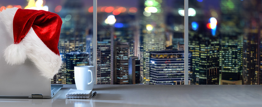诞工作场所全景笔记本电脑桌子上,诞老人帽子办公室,全景夜景的现代市中心摩天大楼商业区图片