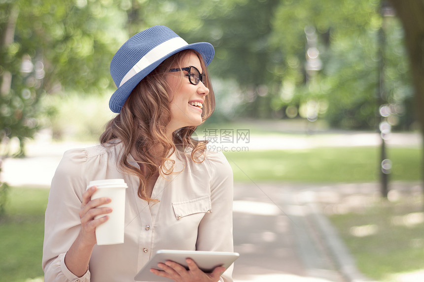 快乐的轻女人着次咖啡杯,手里着平板电脑,喝着咖啡,着夏天的公园背景微笑图片