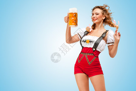 啤酒女郎轻的调情感的瑞士女人穿着红色跳线短裤吊带的传统Dirndl,着蓝色背景的啤酒杯背景