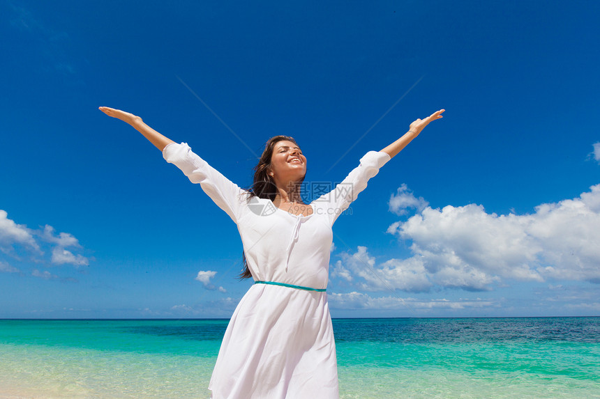 海滩上穿着白色连衣裙的女人穿着白色连衣裙的女人站蓝色的海边背景上,举手图片
