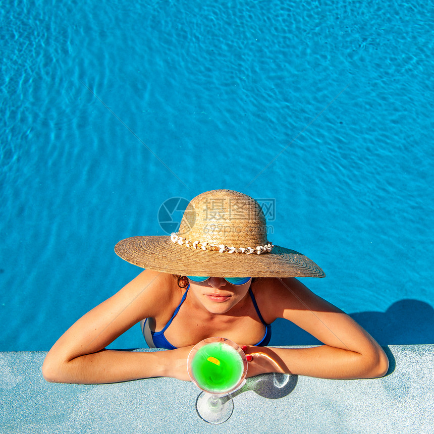 女人池边喝鸡尾酒戴帽子的女人游泳池里用鸡尾酒放松图片