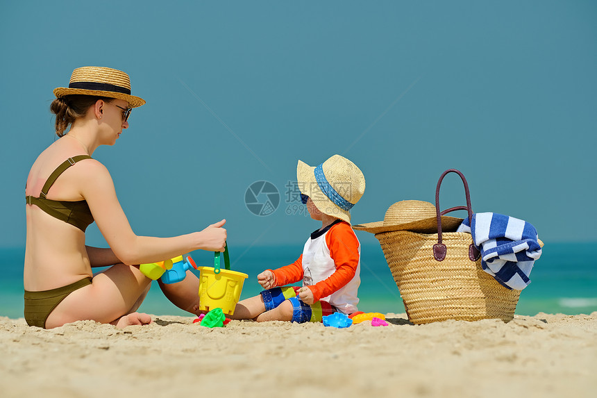 蹒跚学步的男孩海滩上妈妈玩两岁的蹒跚学步的男孩戴着太阳帽妈妈海滩上玩耍图片
