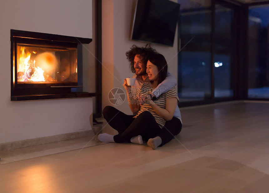 轻的浪漫多民族夫妇坐家里壁炉前的地板上,寒冷的冬夜聊天喝茶图片