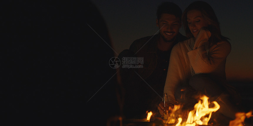 轻的夫妇晚上朋友坐海滩上的篝火旁喝啤酒图片