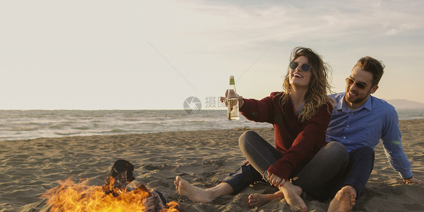 轻夫妇火旁放松,喝啤酒瓶子里的饮料图片
