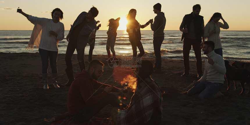 快乐无忧无虑的轻朋友海滩上玩得开心,太阳开始落山的时候篝火旁喝啤酒图片
