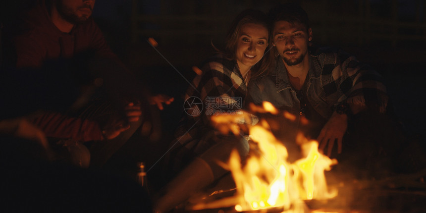 轻的夫妇晚上朋友坐海滩上的篝火旁喝啤酒图片