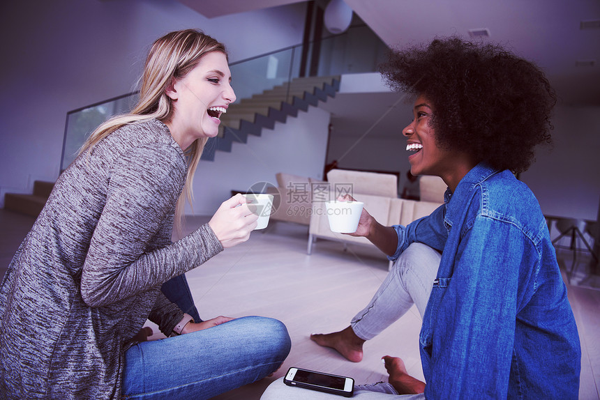 两个轻的笑着的多民族妇女坐地板上,边喝咖啡,边豪华的家里用智能手机喝咖啡图片