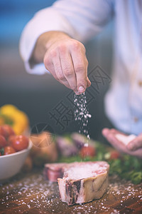 主厨手把盐放多汁的生牛排上,蔬菜放木桌上厨师把盐放多汁的生牛排上背景图片