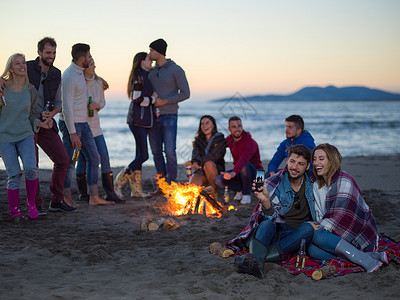 夫妇秋天的海滩聚会上用手机朋友喝啤酒,玩得开心夫妇海滩上朋友享受篝火图片