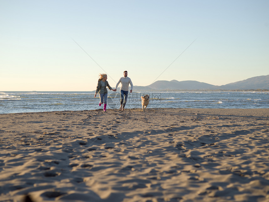 夫妇与狗沙滩上玩得开心自动日Autmun的天,夫妇海滩上跑步,用狗牵着手图片