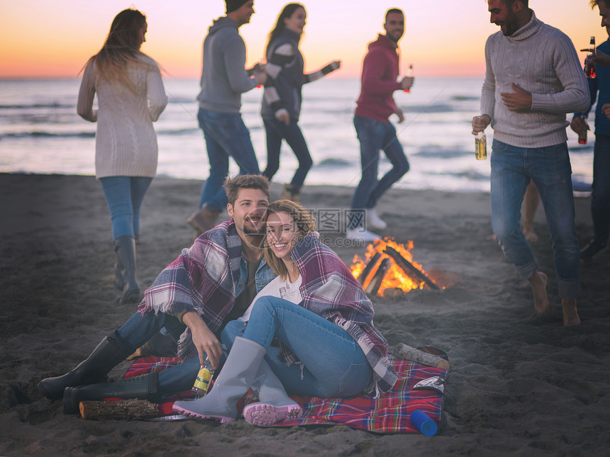夫妇海滩上日落时朋友享受轻的夫妇朋友坐海滩上的篝火旁,日落时喝啤酒彩色过滤器图片