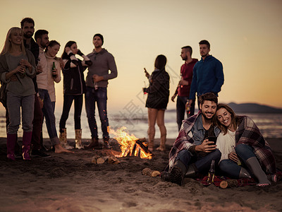 聚是一团火夫妇海滩上朋友享受篝火夫妇海滩聚会上用手机,朋友们喝啤酒,享受趣的彩色过滤器背景