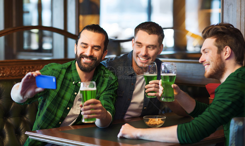 帕特里克节,休闲技术快乐的男朋友喝绿色啤酒,并酒吧酒吧用智能手机自拍棒拍照朋友们酒吧用绿色啤酒自拍图片
