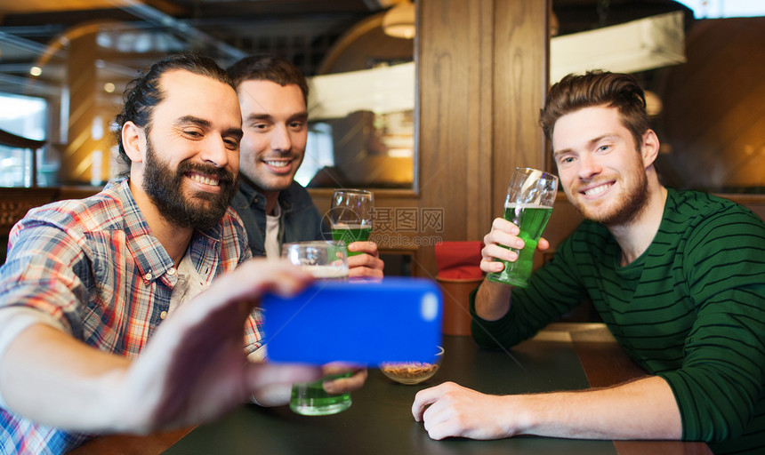 帕特里克节,休闲技术快乐的男朋友喝绿色啤酒,并酒吧酒吧用智能手机自拍棒拍照朋友们酒吧用绿色啤酒自拍图片