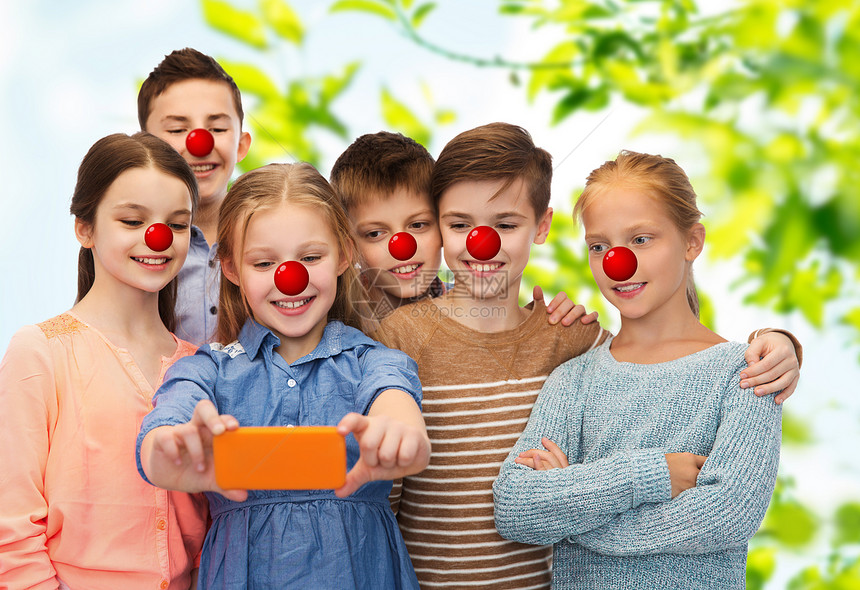 红鼻子日,慈善童的快乐的孩子与小丑鼻子自拍智能手机绿色的自然背景孩子们红鼻子日用智能手机自拍图片