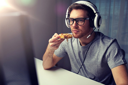 披萨现实的技术,游戏,娱乐活动,让们的游戏人的轻人耳机与个人电脑吃比萨饼,同时家里玩游戏流媒体播放演练视频戴耳机的男人背景