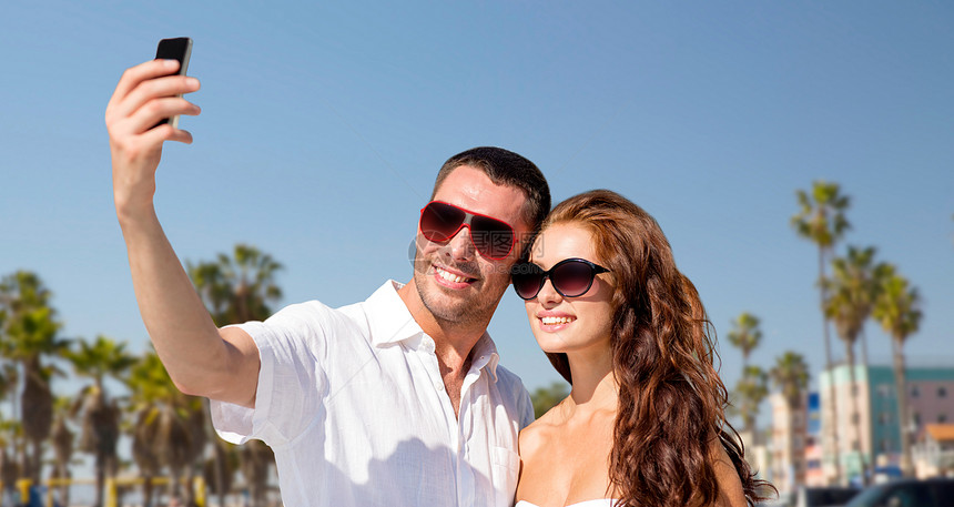 旅行,旅游技术微笑夫妇戴着太阳镜,加州威尼斯海滩背景上用智能手机自拍戴着墨镜的夫妇威尼斯海滩上自拍图片