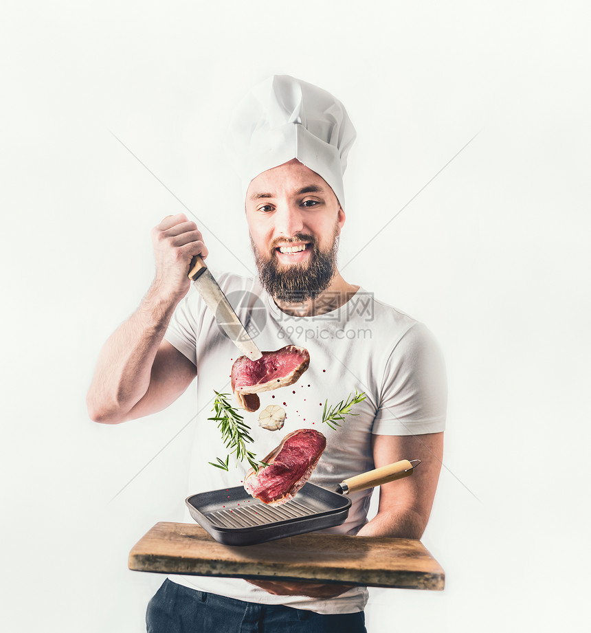 轻开朗的厨师,留着胡子,着个煎锅,牛排刀子,灯光的背景下空中飞舞肉类食品烹饪图片