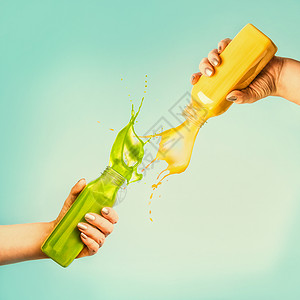 女手着黄色绿色的瓶子,蓝色背景上溅上冰沙果汁,热带树叶水果夏季饮料的图片