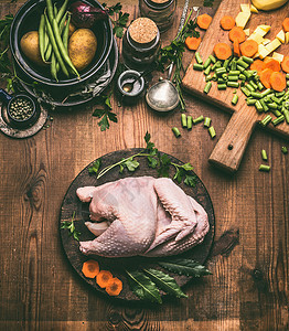 原始整鸡乡村木制厨房桌子背景与蔬菜,烹饪勺子砧板,顶部视图鸡肉烹饪准备图片
