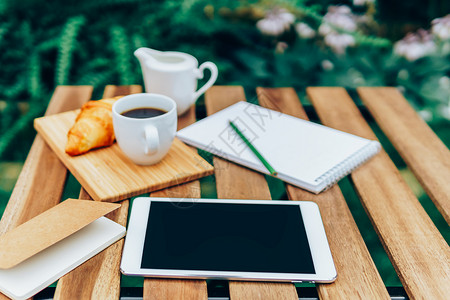 早上早餐绿色花园与法国牛角包,咖啡杯,橙汁,平板电脑笔记簿木桌上背景图片