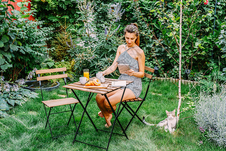 轻而魅力的女人绿色花园里吃早餐,法国牛角包甜甜圈咖啡杯橙汁平板电脑木制桌子上的笔记簿背景图片