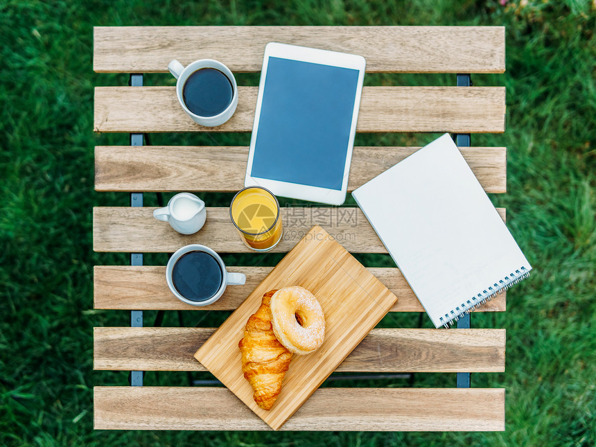 早上早餐绿色花园与法国牛角包,甜甜圈,咖啡杯,橙汁,平板电脑笔记簿木桌上图片